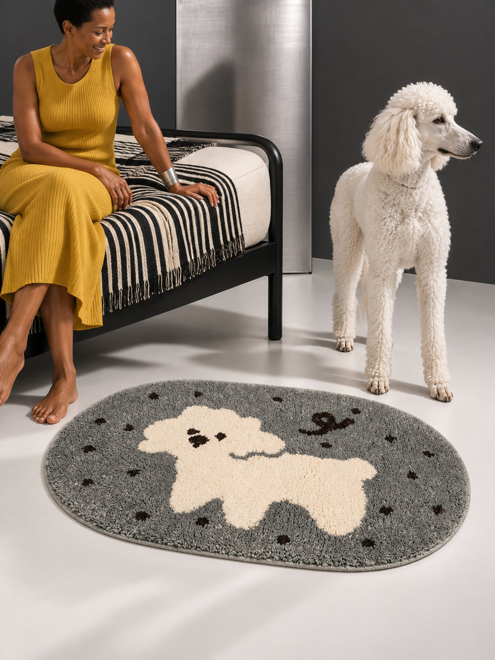 Gray polka-dot pup oval mat, 50 x 80 cm, beside a seated person, a white poodle and a striped bed.
